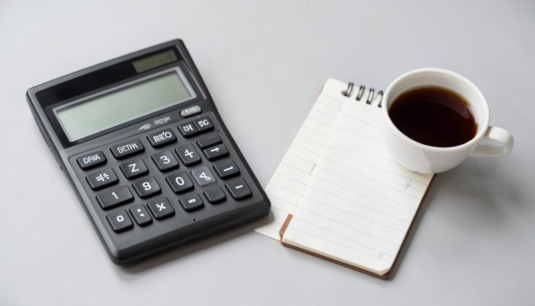 A clean desktop workspace with a calculator, a notepad, and a cup of coffee on a light grey surface